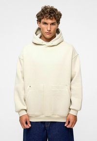 Beige hoodie van zacht materiaal, met een grote capuchon, twee voorzakken en ribgebreide manchetten. Casual, oversized pasvorm met minimale details.