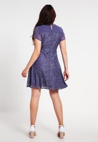 Robe en dentelle violette avec des manches courtes et transparentes, jupe évasée, détails à la taille et découpe en forme de clé dans le dos. Portée avec des sandales à talons blocs blanches.