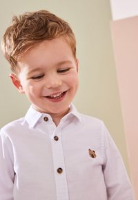 Camicia bianca con bottoni marroni e un piccolo ricamo di un orso marrone sul petto. Tessuto liscio su uno sfondo morbido.