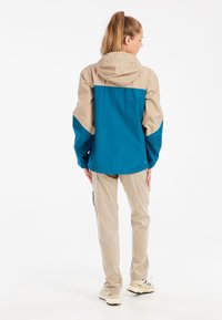 Nepremokavá bunda v odtieni teal a béžovej, s kapucňou, dlhými rukávmi a kontrastnými bočnými panelmi. V kombinácii s béžovými nohavicami a teniskami.