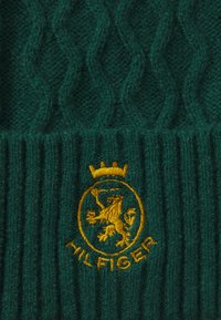 Zielona dziana czapka typu beanie z fakturowanym wzorem w warkocze, ściągaczem i haftowanym złotym herbem lwa z koroną oraz napisem „HIL FIGER”.