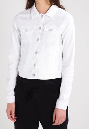 Vrouw die een nauwsluitend wit jasje met knopen en lange mouwen draagt, en een zwarte broek tegen een effen achtergrond.