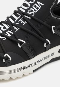 Juodas sportinis batelis su elastinga viršutine dalimi, baltais logotipais ir kontrastingomis raištelėmis, bei tekstūruotu baltu padu su prekiniu ženklu.