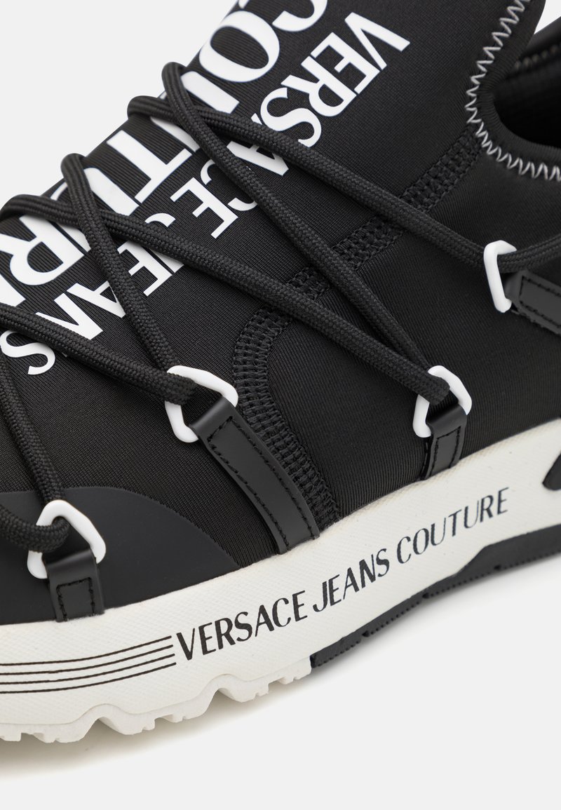 Schwarzer Sportschuh mit einem elastischen Obermaterial, weißen Logos und kontrastierenden Schnürsenkeln sowie einer strukturierten weißen Sohle mit Markenlogo.