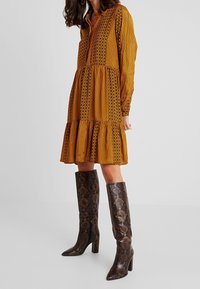 Langärmliges, knöpfbares Kleid in Senfgelb mit vertikalen Streifen und Blumenmustern. Kombiniert mit kniehohen braunen Stiefeln im Schlangenmuster.