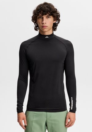 Młody mężczyzna z kręconymi włosami, ubrany w dopasowany czarny długi rękaw z logo J.LINDEBERG na kołnierzu i rękawie oraz jasne zielone spodnie.