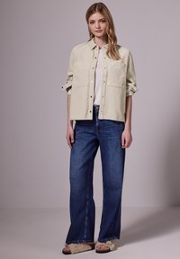 Chemise en velours côtelé vert clair avec deux poches devant, associée à un jean large bleu et des sandales à sliders beige en fourrure. Tenue neutre et décontractée.