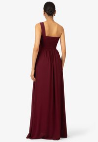 Robe maxi sans manches bordeaux avec un corsage ajusté en smocks et une jupe fluide, dotée d'une seule bretelle et d'une texture de tissu lisse.