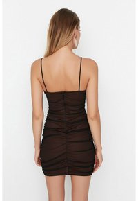 Femme aux longs cheveux portant une mini-robe noire ajustée et froncée avec des bretelles fines, debout de dos sur un fond blanc uni.