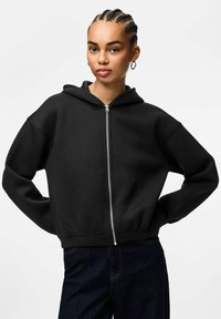 Felpa con zip nera, con tasca frontale, vestibilità oversize e orlo a coste. Presenta un cappuccio con coulisse e un tessuto morbido e strutturato.