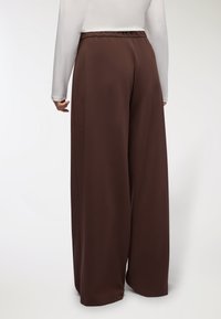 Brązowe, szerokie spodnie z elastyczną talią, charakteryzujące się gładką fakturą i opadającym krojem, w zestawieniu z białą bluzką z długim rękawem.