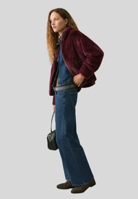 Jachetă din blană artificială burgundy peste cămașă din denim, asociată cu blugi evazați albaștri și cizme maro. O geantă de mână neagră completează ținuta.
