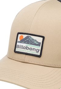 Bézs színű sapka strapabíró anyagból, téglalap alakú folttal, amelyen hegyi grafika, nap és a "Billabong Adventure Division" szöveg található.