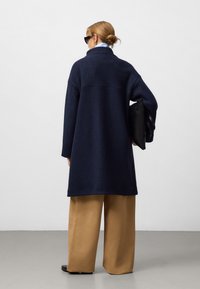 Manteau en mélange de laine navy avec un col haut, une coupe oversize et de longues manches, associé à un pantalon ample beige et une pochette noire.