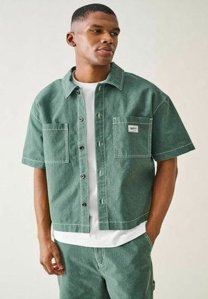 Młody mężczyzna ubrany w zieloną jeansową krótką kurtkę i pasujące spodnie na białej koszulce, stojący z jedną ręką w kieszeni.