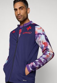 Lila sportdzseki cipzáras záródással, virágmintás ujjakkal, piros logó részletekkel és zsebekkel. Sima textúrájú, modern kialakítás.