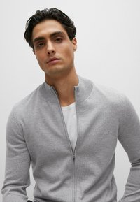 Maglione grigio con zip e colletto alto, polsini a coste e un logo discreto sul petto, indossato sopra una T-shirt bianca. Texture liscia.