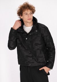 Schwarze Tarnjacke mit hohem Kragen, Druckknöpfen und legerem Schnitt. Hergestellt aus einem glatten, strapazierfähigen Material. Mit Taschen akzentuiert.