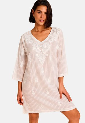 Femme aux cheveux courts et foncés portant une robe blanche brodée de fleurs avec un col en V et des manches trois-quarts, debout sur un fond uni.
