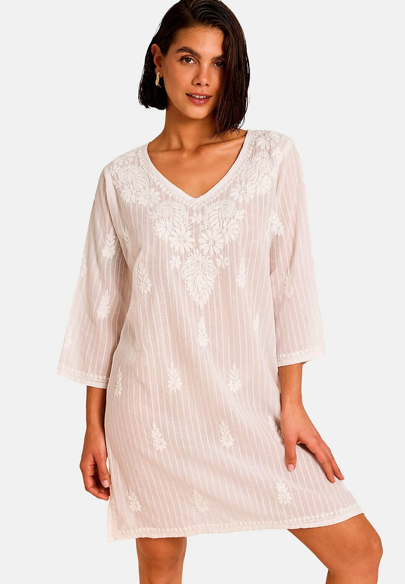 Femme aux cheveux courts et foncés portant une robe blanche brodée de fleurs avec un col en V et des manches trois-quarts, debout sur un fond uni.