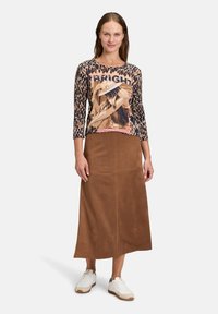 Brązowa zamszowa długa spódnica połączona z wzorzystą bluzką z nadrukiem zwierzęcym i sztuką graficzną. Modelka w białych sneakersach.