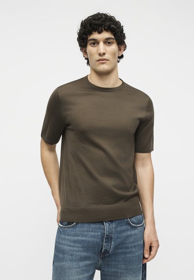 Νέος άνδρας με σγουρά μαλλιά που φοράει ένα καφέ εφαρμοστό κοντομάνικο πουκάμισο και μπλε τζιν, στέκεται με ουδέτερη έκφραση μπροστά σε λευκό φόντο.