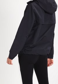 Black lightweight jacket with a hood, textured fabric, adjustable cuffs, and a relaxed fit from shoulder to hem. Features a back flap detail.