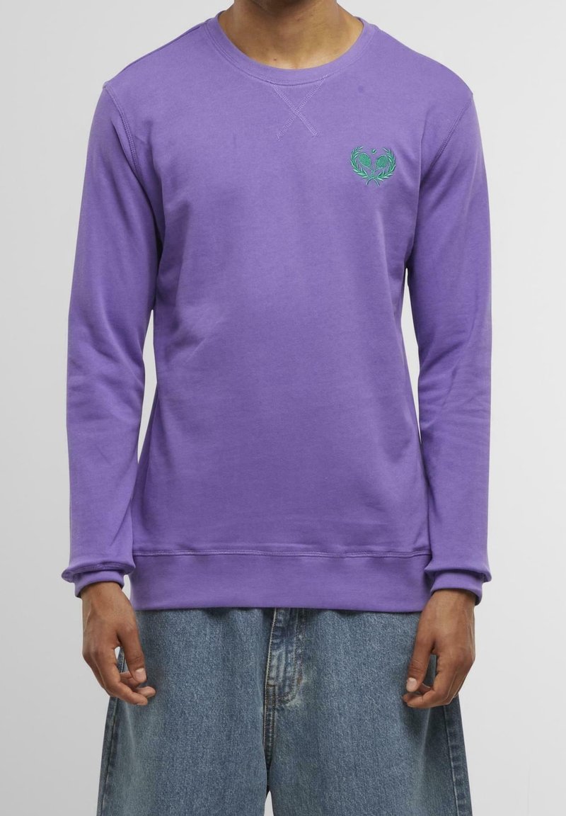 Homme portant un sweat-shirt violet à manches longues et col rond avec un petit logo vert brodé sur la poitrine et un jean bleu ample.