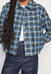 Blau- und olivfarbener karierter, verkürzter Jacke mit großen Knöpfen und fransigen Rändern. Strukturierter Stoff mit sichtbarem Webmuster. Kombiniert mit dunkler Jeans.