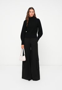 Schwarzer Strickpullover mit hohem Kragen, kombiniert mit weit geschnittenen schwarzen Hosen mit Cargo-Taschen, abgerundet mit einer kleinen rosa Handtasche.