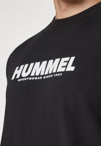 Svart hettegenser med rund hals, med fet hvit tekst "HUMMEL SPORTSWEAR SINCE 1923." Glatt tekstur, minimalistisk design.