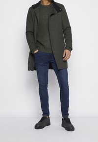 Manteau long vert avec col noir, pull vert texturé, jean bleu et baskets noires. Coupe slim avec un design moderne.
