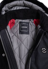 Chaqueta negra con forro acolchado a cuadros en blanco y negro, acentos de tela roja, cremalleras de metal y etiquetas de la marca IKKS tanto en el interior como en el exterior.