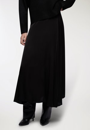 Gonna maxi in satin nero con una silhouette fluida, caratterizzata da un alto girovita e una superficie liscia. Indossata con stivaletti aderenti in pelle nera.