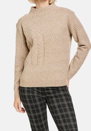 Pull beige en maille torsadée avec motifs texturés associé à un pantalon à carreaux noir et blanc sur une personne debout.