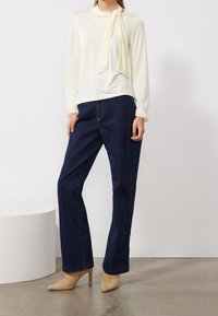 Mujer con una blusa de manga larga color crema con tiras en los hombros, jeans rectos de mezclilla azul oscuro y botas de tacón alto de punta beige, de pie en el interior.