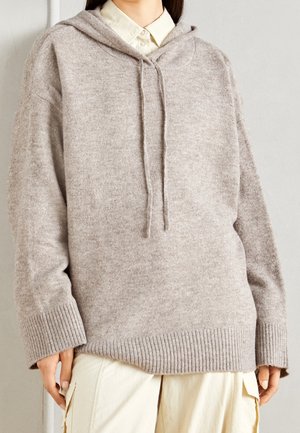 Hellbrauner übergroßer Kapuzenpullover aus weichem Stoff, mit Kordelzug, gerippten Bündchen und Saum, getragen über einem Hemd mit Kragen.