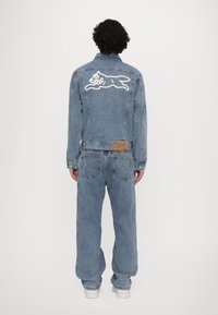 Giubbotto di jeans con grafica nella parte posteriore di un animale che salta, colore blu sbiadito, design classico e texture leggera; abbinato a pantaloni di jeans a vestibilità larga.