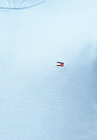 Világos kék kötött pulóver, amelyen egy kis téglalap alakú logó található fehér, piros és fekete csíkokkal a mellrészen.