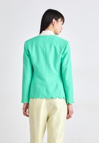 Mintgroene getailleerde blazer met geschulpte zoomdetails, gecombineerd met glanzende gouden broeken, met lange mouwen en een gestructureerde pasvorm.