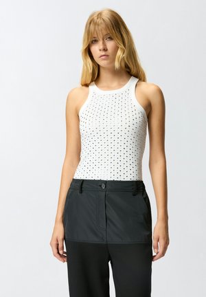 Witte geribde tanktop met zwarte stippenpatronen, gecombineerd met donkere stoffen broek en een knoopsluiting. Eenvoudige, sportieve snit.