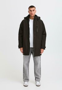 Svart lang jakke med hette, glidelås og framlommer. Båret over en hvit skjorte og kombinert med lysegrå jeans og joggesko.