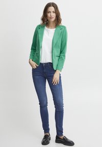 Zelený blazer s klopami, bílá košile, modré úzké džíny a černé mokasíny. Hladká tkanina, přiléhavý design a dva přední kapsy na blazeru.