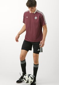 Maillot rayé bordeaux avec un col blanc et un logo, associé à un short noir et des chaussettes noires rayées, se tenant à côté d'un ballon de soccer noir et blanc.