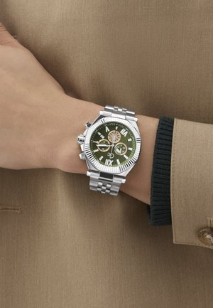 Ezüst rozsdamentes acél óra zöld számlappal, római számokkal, három segédszámlálóval és textúrázott kerettel. Egy kézen viselve, bézs szövet előtt.