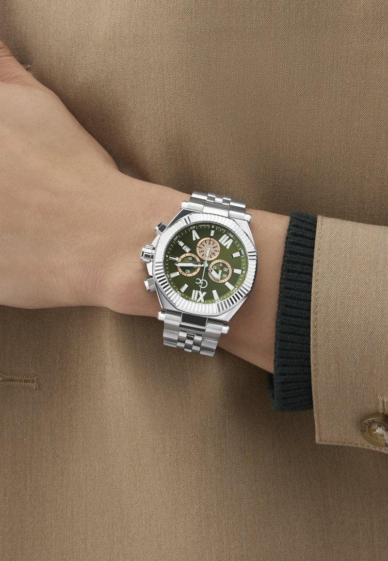 Srebrna ura iz nerjavečega jekla z zelenim številčnico, rimskimi številkami, tremi malimi številčnicami in teksturiranim robom. Nosimo jo na roki ob bež tkanini.