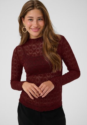 Jeune femme souriante, portant un haut en dentelle bordeaux à manches longues et de grandes boucles d'oreilles dorées en forme d'anneau, debout devant un fond clair uni.