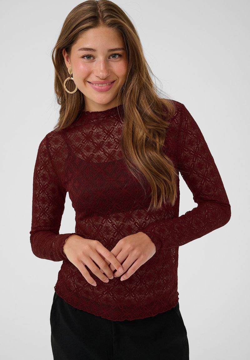 Jeune femme souriante, portant un haut en dentelle bordeaux à manches longues et de grandes boucles d'oreilles dorées en forme d'anneau, debout devant un fond clair uni.