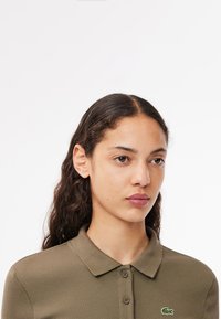 Olivgrön ribbad pikétröja med krage, som har två knappar och en liten broderad logotyp på vänster sida. Slät tygstruktur.