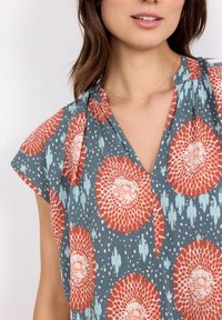 Blouse à manches courtes en tissu bleu avec un motif floral rouge et blanc, présentant un décolleté en V et un matériau texturé léger.